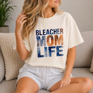 Könnte beinhalten: Cremefarbenes T-Shirt mit dem Schriftzug "BLEACHER MOM LIFE" in blauen und orangefarbenen Aquarellbuchstaben. Das Shirt ist ein lässiger Stil mit kurzen Ärmeln.