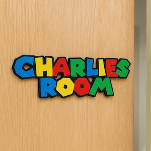 Op de afbeelding: Een kleurrijk bord met de tekst "CHARLIES ROOM" in een speels, blokkerig lettertype. De letters zijn zwart omlijnd en gevuld met blauw, geel, rood en groen. Het bord is bevestigd op een lichtgekleurde houten deur.