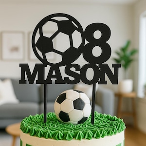 May include: A soccer-themed birthday cake with green and white stripes and a soccer ball decoration. The cake is topped with a black cake topper that reads "08 MASON" and a soccer ball.