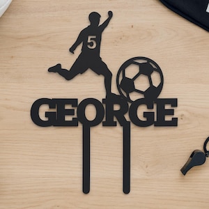 May include: Black cake topper featuring a soccer player kicking a ball, with the name "GEORGE" below. The player has the number 5 on their jersey. A soccer ball, cleats, and a whistle are also visible.