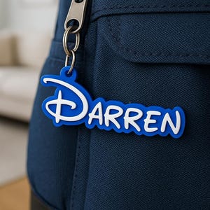 Könnte beinhalten: Ein blau-weißer Schlüsselanhänger mit dem Namen "Darren" in einer stilisierten Schriftart, befestigt an einem silberfarbenen Metallverschluss. Der Schlüsselanhänger hängt an einem marineblauen Rucksack.