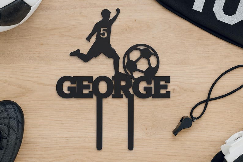 May include: Black cake topper featuring a soccer player kicking a ball, with the name "GEORGE" below. The player has the number 5 on their jersey. A soccer ball, cleats, and a whistle are also visible.