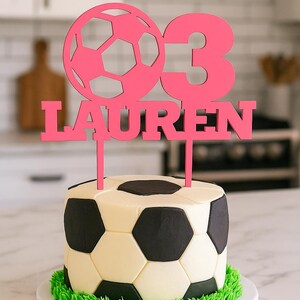 May include: A soccer ball-themed cake with a pink topper that reads "03 LAUREN." The cake is decorated to resemble a soccer ball with black and white panels and green frosting around the base. The cake sits on a white plate.
