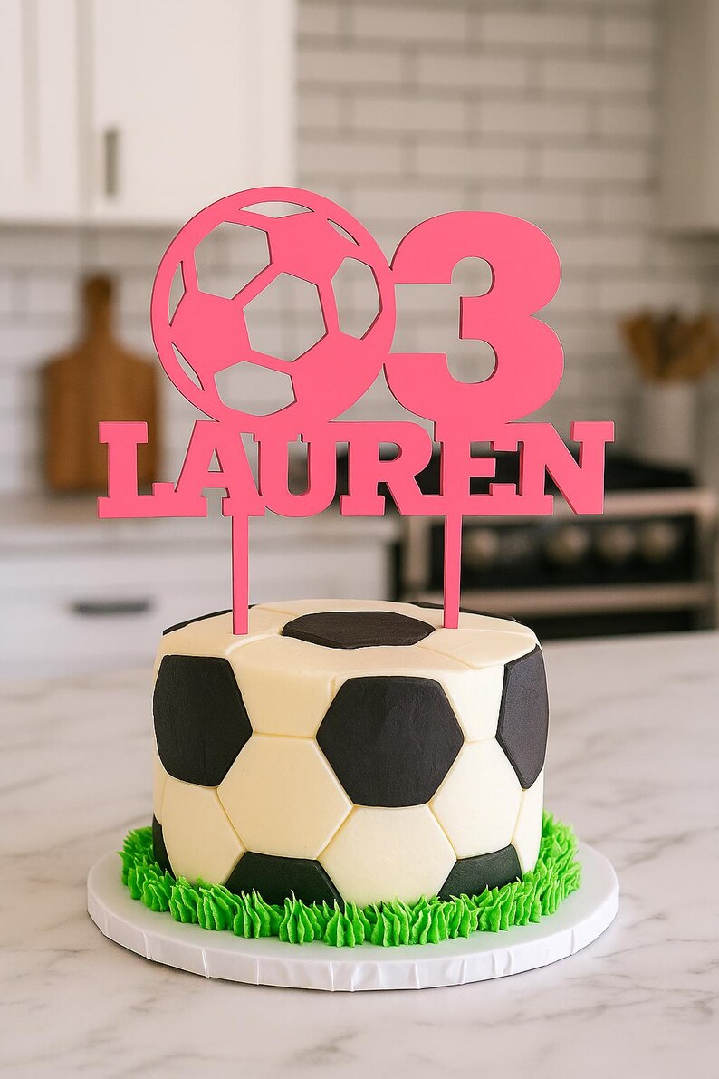 May include: A soccer ball-themed cake with a pink topper that reads "03 LAUREN." The cake is decorated to resemble a soccer ball with black and white panels and green frosting around the base. The cake sits on a white plate.