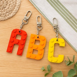 Könnte beinhalten: Drei Schlüsselanhänger mit den Buchstaben "ABC". Jeder Buchstabe hat eine andere Farbe: rot, orange und gelb. Die Buchstaben sind als Bausteine gestaltet, mit silberfarbenen Verschlüssen.