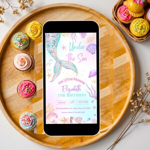 K&ouml;nnte beinhalten: Ein Smartphone zeigt eine Einladung zum Geburtstag mit Meerjungfrauen-Motiv und dem Text "Under the Sea". Die Einladung zeigt eine Meerjungfrauenschwanz, Muscheln und den Namen "Elizabeth". Cupcakes mit farbenfrohem Zuckerguss und Streuseln umgeben das Telefon auf einem Holztablett.