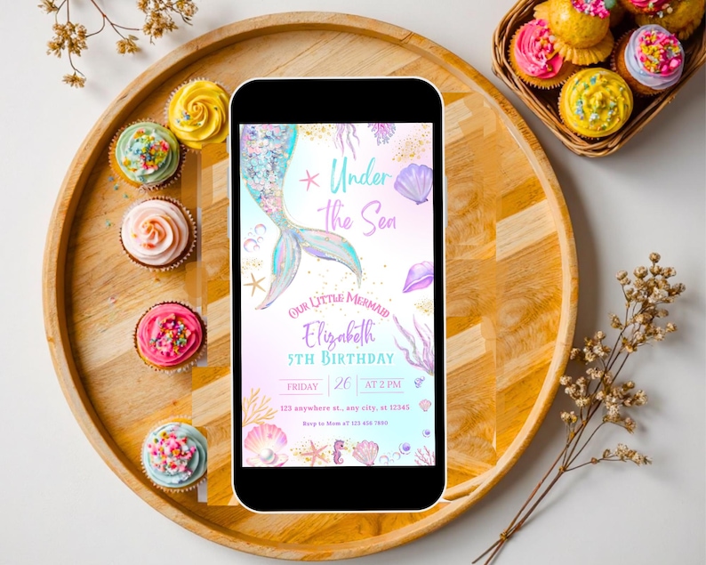 K&ouml;nnte beinhalten: Ein Smartphone zeigt eine Einladung zum Geburtstag mit Meerjungfrauen-Motiv und dem Text "Under the Sea". Die Einladung zeigt eine Meerjungfrauenschwanz, Muscheln und den Namen "Elizabeth". Cupcakes mit farbenfrohem Zuckerguss und Streuseln umgeben das Telefon auf einem Holztablett.