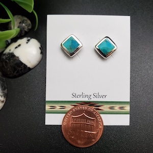 May include: A pair of square-shaped sterling silver stud earrings with turquoise centers. The earrings are displayed on a white card with the words "Sterling Silver" printed on it. A penny is placed below the card.