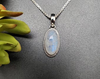 May include: A silver necklace featuring an oval moonstone pendant. The pendant has a silver rope-style border and a light blue and white stone. The necklace is on a dark grey surface.