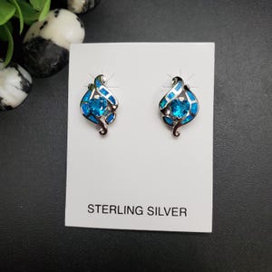 May include: A pair of sterling silver earrings with a blue gemstone center and blue opal accents. The earrings are displayed on a white card with the text "STERLING SILVER". The earrings have a swirling design.