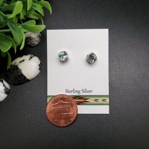 May include: A pair of sterling silver stud earrings with square, light blue gemstones. The earrings are displayed on a white card with the text "Sterling Silver". A penny is next to the earrings.