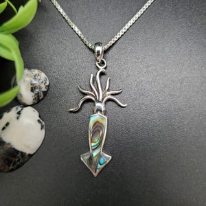 Op de afbeelding: Een zilverkleurige hangerketting met een inktvisontwerp. De hanger heeft een centrale abalone schelpinleg met iriserende kleuren blauw, groen en goud. De tentakels van de inktvis strekken zich uit en de ketting is van zilver.