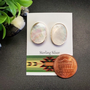 May include: A pair of oval-shaped mother-of-pearl earrings with gold-colored trim. The earrings are displayed on a white card with the text "Sterling Silver" and a Native American-inspired design. A penny is next to the earrings.