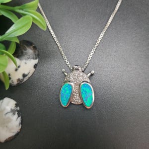 May include: A silver-toned necklace featuring a beetle-shaped pendant. The insect's body is adorned with two vibrant, iridescent blue-green oval stones for wings and a head section encrusted with small, clear, sparkling white stones.
