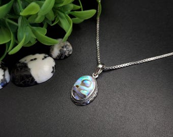 Pode incluir: Um colar de prata com um pingente oval com uma concha de abalone iridescente. O pingente é emoldurado por uma borda de prata decorativa. O colar é exibido em uma superfície preta com folhagem verde ao fundo.