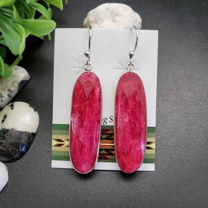 May include: A pair of dangle earrings featuring large, oval-shaped, vibrant pink gemstones set in silver-colored metal. The earrings hang from simple silver hooks. The gemstones have a faceted appearance, catching the light. The earrings are displayed on a white card.