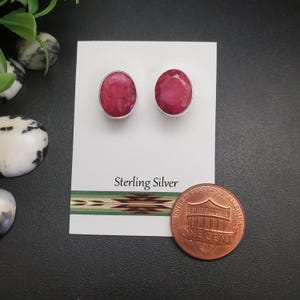 May include: A pair of sterling silver stud earrings with oval-shaped, deep red gemstones. The earrings are displayed on a white card with "Sterling Silver" printed on it. A penny is placed next to the earrings for size comparison.