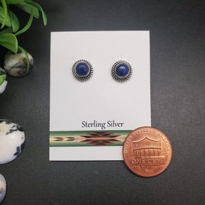 May include: Sterling silver stud earrings with a round, dark blue stone center and a twisted rope design border. The earrings are displayed on a white card with the words "Sterling Silver" printed on it. A penny is next to the earrings.