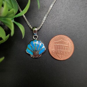 May include: A silver-toned necklace featuring a seashell-shaped pendant inlaid with bright blue opal-like material. The pendant is positioned next to a United States one-cent coin for scale, showing the relative size of the jewelry piece.