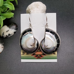 May include: A pair of silver earrings with a shell-like design. Each earring features a pearlescent, iridescent center framed by a black border and a silver edge. The earrings hang from silver hooks, showcasing a unique, naturalistic aesthetic.