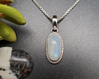 May include: A silver necklace with an oval moonstone pendant. The pendant is framed by a twisted rope design. The moonstone has a pearly, iridescent sheen. The chain is a delicate box chain. The background is dark.