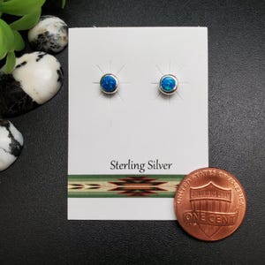 May include: A pair of sterling silver stud earrings with round, blue opal stones. The earrings are displayed on a white card with the text "Sterling Silver" and a decorative green and brown pattern. A US one-cent coin is next to the earrings.