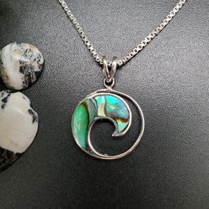 May include: A silver pendant necklace featuring an abalone shell design in shades of green, blue, and brown. The pendant is set in a silver swirl design and hangs from a silver chain. Two decorative stones are in the background.