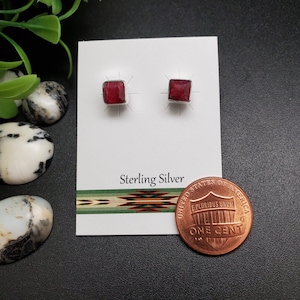 May include: A pair of square, red gemstone stud earrings set in sterling silver. The earrings are displayed on a white card with the text "Sterling Silver". A penny is next to the earrings for size reference.