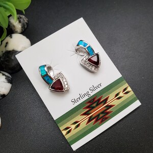 May include: A pair of sterling silver earrings. Each earring features a triangular, deep red gemstone, surrounded by small clear stones. The top of each earring has a curved section with blue-green inlay. The card reads "Sterling Silver".