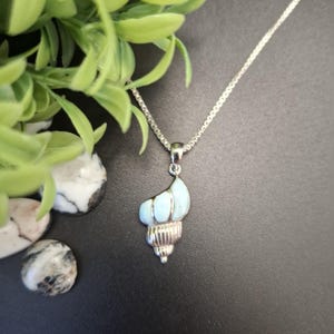 May include: A silver necklace featuring a pendant shaped like a seashell. The pendant has a polished silver base with a smooth, light blue stone inlay that mimics the spiral form of a shell. The chain is a thin, metallic silver box link style.