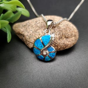 May include: A silver-toned pendant shaped like a nautilus shell, featuring vibrant blue opal inlays segmented by polished metal lines. The pendant hangs from a delicate silver-toned box chain, showcasing a spiral design with a reflective finish.