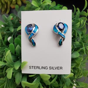May include: A pair of sterling silver earrings with a swirling design. Each earring features a dark blue gemstone at the center, surrounded by a vibrant blue and turquoise inlay. The text "STERLING SILVER" is printed on the display card.