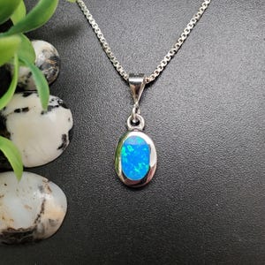 May include: A silver necklace with an oval pendant featuring a vibrant blue opal gemstone. The opal is set in a silver bezel and hangs from a delicate silver chain. The background is a dark, textured surface.