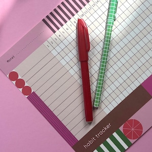 Könnte beinhalten: Ein Habit-Tracker-Blatt mit einem Raster, einem roten Stift, einem grünen Stift mit Karomuster, einem rot-weiß karierten Federmäppchen und zwei Klebebandrollen. Der Tracker enthält die Aufschriften "Monat" und "habit tracker" sowie geometrische Formen.