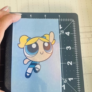 May include: A rectangular card featuring a cartoon illustration of a blonde character with blue eyes, wearing a blue dress with a black stripe, positioned against a light blue background. The card measures approximately 9 cm by 12.7 cm.