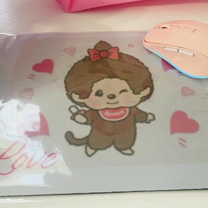 Może przedstawiać: Jasnoróżowa bezprzewodowa mysz komputerowa spoczywa na podkładce pod mysz z postacią z kreskówki z czerwoną kokardą i śliniaczkiem. Podkładka ma jasnoniebieską obwódkę, różowe serca i słowo "Love" w różowym skrypcie.