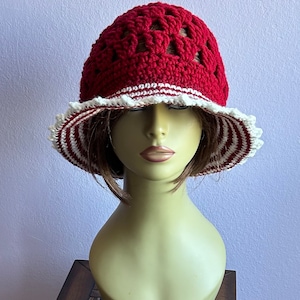 May include: A red crocheted sun hat with white trim and a scalloped edge. The hat is on a mannequin head.