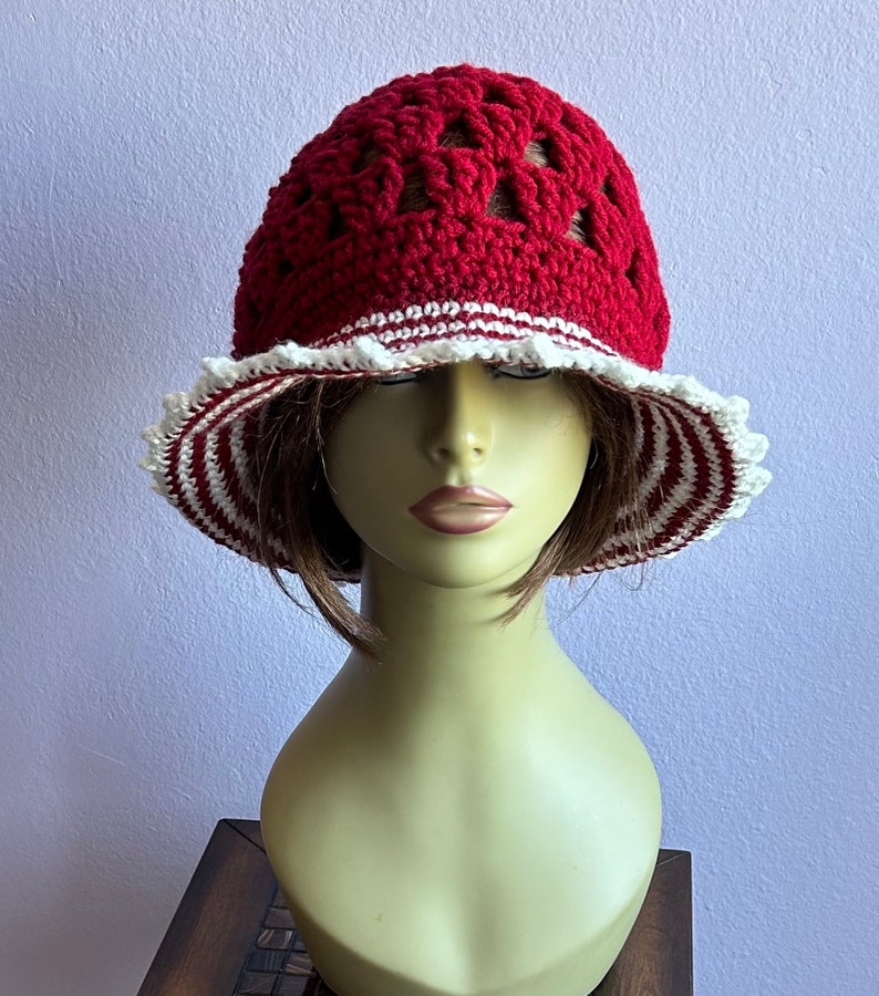 May include: A red crocheted sun hat with white trim and a scalloped edge. The hat is on a mannequin head.