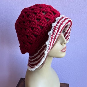 May include: A red crocheted hat with a white and red striped brim. The hat is on a mannequin head.