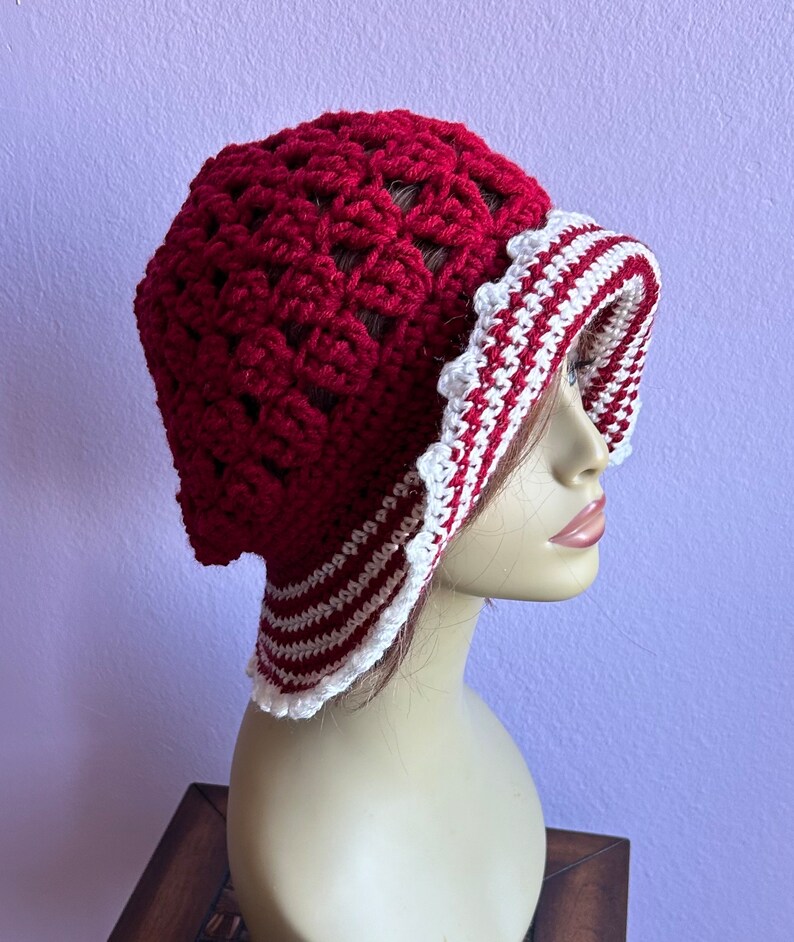 May include: A red crocheted hat with a white and red striped brim. The hat is on a mannequin head.