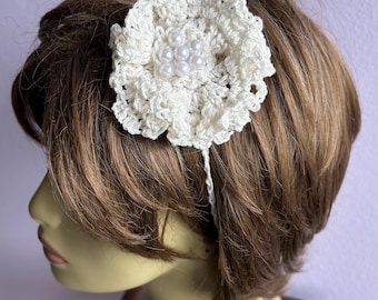 May include: A cream-colored, crocheted flower hair accessory with a cluster of faux pearls at the center. The flower is attached to a thin, white headband. The accessory is worn on a mannequin with short, brown hair.