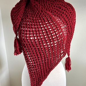 May include: A deep red, hand-knitted shawl with a triangular shape. The shawl features a textured open weave design and is adorned with three tassels at the points. The item is displayed on a mannequin.