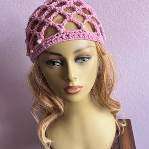 May include: A light pink crocheted hat with a net-like design and small white beads. The hat is worn on a mannequin head with blonde hair and a neutral expression. The background is a solid light purple.