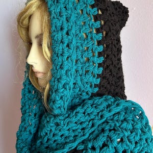 May include: A handmade crochet hood in teal and black. The hood is designed with a cowl neck and a hood that covers the head. The teal sections have an open, lacy stitch, while the black sections are solid. The hood is displayed on a mannequin.