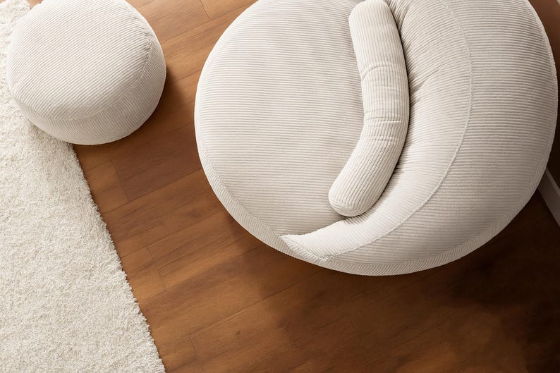 Puede incluir: Vista a&eacute;rea de dos muebles tapizados en pana color crema. Uno es un otom&aacute;n redondo, el otro una pieza m&aacute;s grande y redondeada con un coj&iacute;n curvo. Se encuentran sobre un suelo de madera junto a una alfombra blanca.