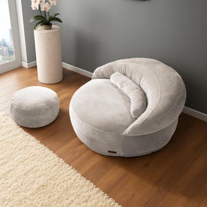Puede incluir: Un sill&oacute;n puf de pana gris claro con un otom&aacute;n a juego. La silla tiene respaldo curvo y un coj&iacute;n cil&iacute;ndrico. Una alfombra color crema y una peque&ntilde;a pintura tambi&eacute;n est&aacute;n en la habitaci&oacute;n. La silla est&aacute; dise&ntilde;ada para la relajaci&oacute;n.