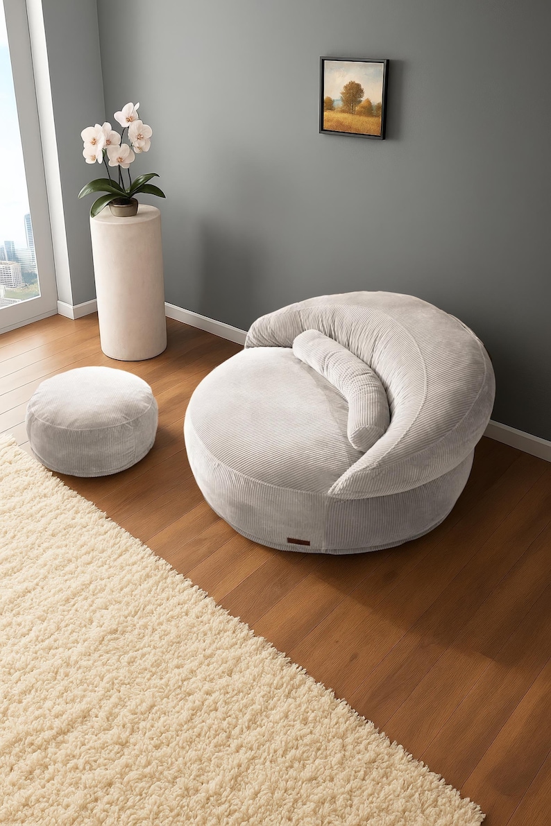 Puede incluir: Un sill&oacute;n puf de pana gris claro con un otom&aacute;n a juego. La silla tiene respaldo curvo y un coj&iacute;n cil&iacute;ndrico. Una alfombra color crema y una peque&ntilde;a pintura tambi&eacute;n est&aacute;n en la habitaci&oacute;n. La silla est&aacute; dise&ntilde;ada para la relajaci&oacute;n.