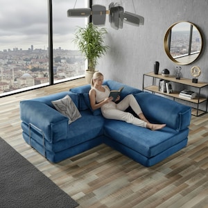 Op de afbeelding: Een blauwe hoekbank van corduroy met een decoratief kussen. De bank heeft een metalen frame en staat in een moderne woonkamer met een groot raam met uitzicht op een stad.