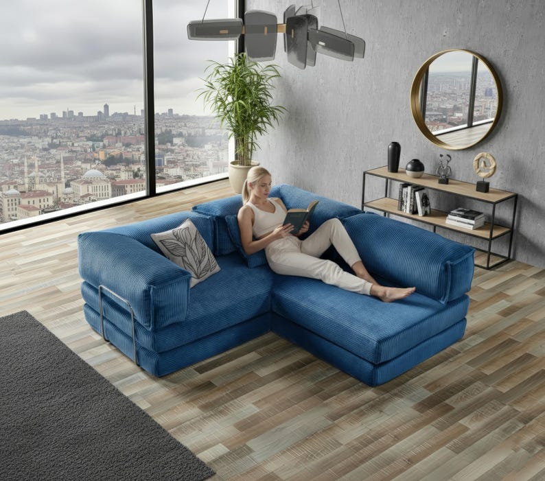 Op de afbeelding: Een blauwe hoekbank van corduroy met een decoratief kussen. De bank heeft een metalen frame en staat in een moderne woonkamer met een groot raam met uitzicht op een stad.