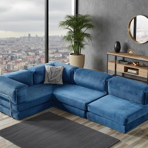 Op de afbeelding: Een grote, modulaire blauwe corduroy hoekbank. De bank heeft een hoekconfiguratie en een decoratief kussen met een bladerenpatroon. De bank staat op een donkergrijs tapijt, met uitzicht op de stad door een groot raam.